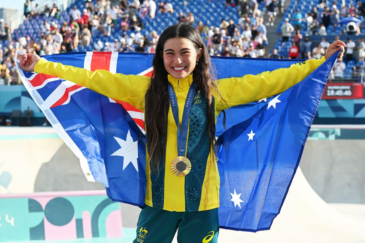 A girl wearing a green and gold tracksuit and with a gold medal around her neck holds up an Australian flag behind her