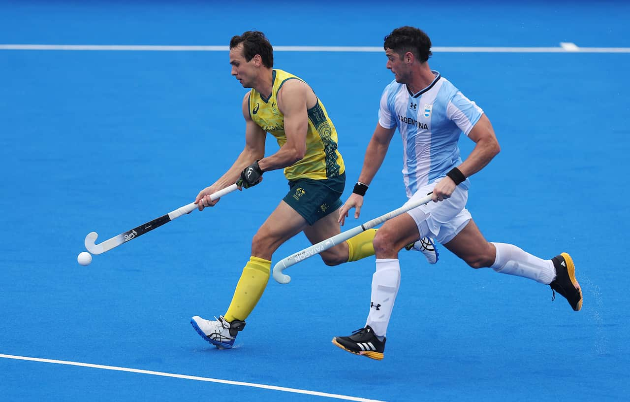 Two men on a hockey field. One is wearing a green and gold uniform and the other a white and blue uniform. 