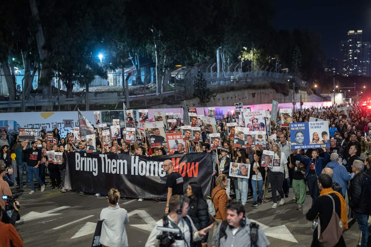 A group of protesters march through a street. Those at the front carry a large banner reading "Bring Them Home Now". 