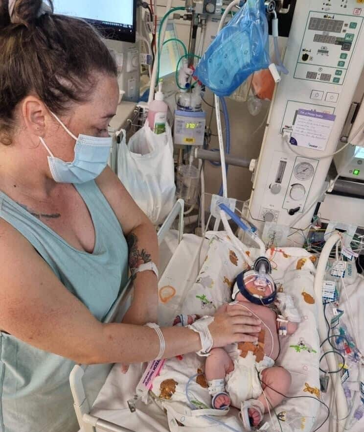 A woman wearing a mask places her hand on a baby in a hospital crib