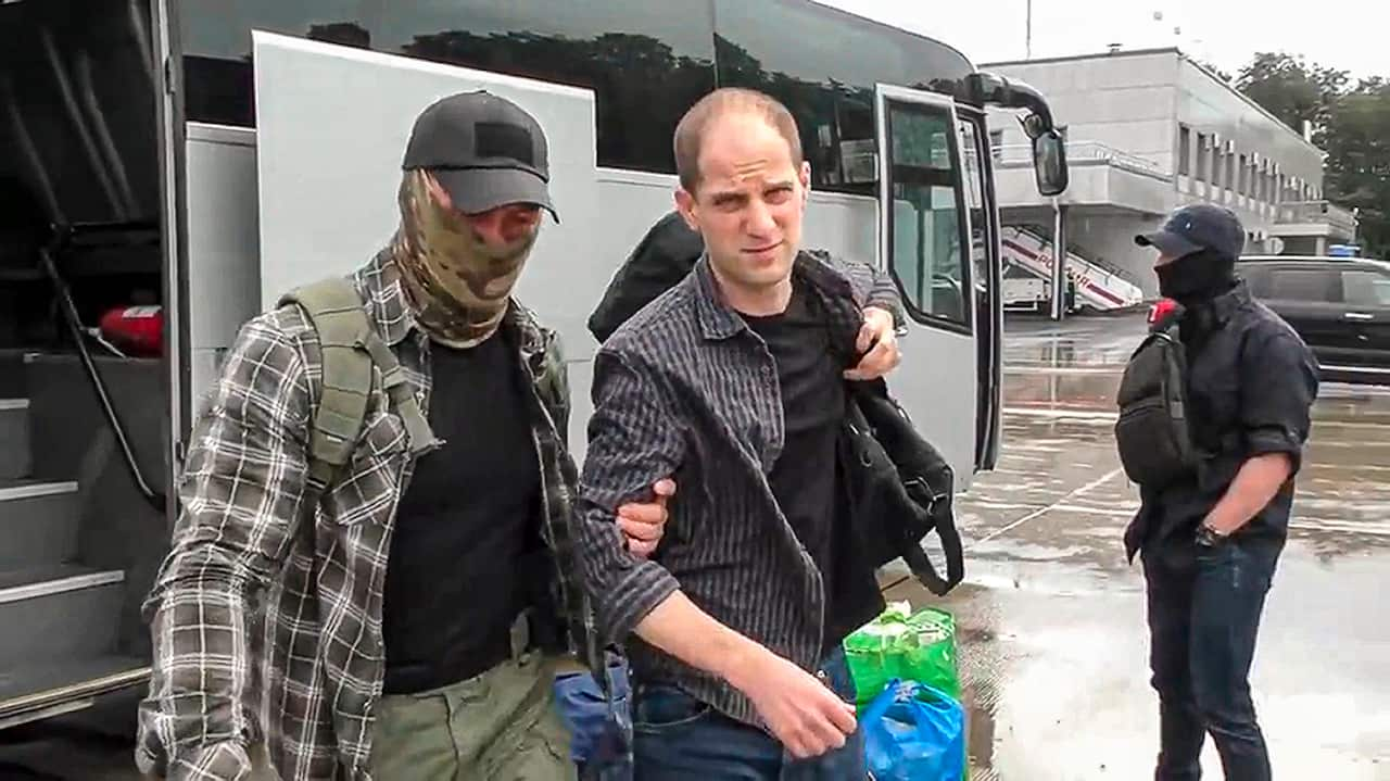 A man wearing a black T-shirt and chequered collared shirt is led by the arm by a man with his face covered by a military camouflage face mask. 