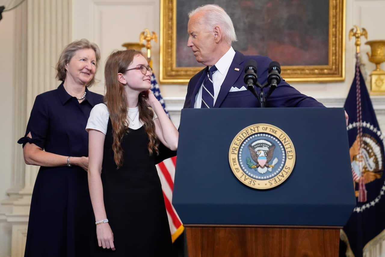 A teenage girl smiling next to US President Joe Biden, who is standing behind a lectern with an official presidential coat of arms on the front. 