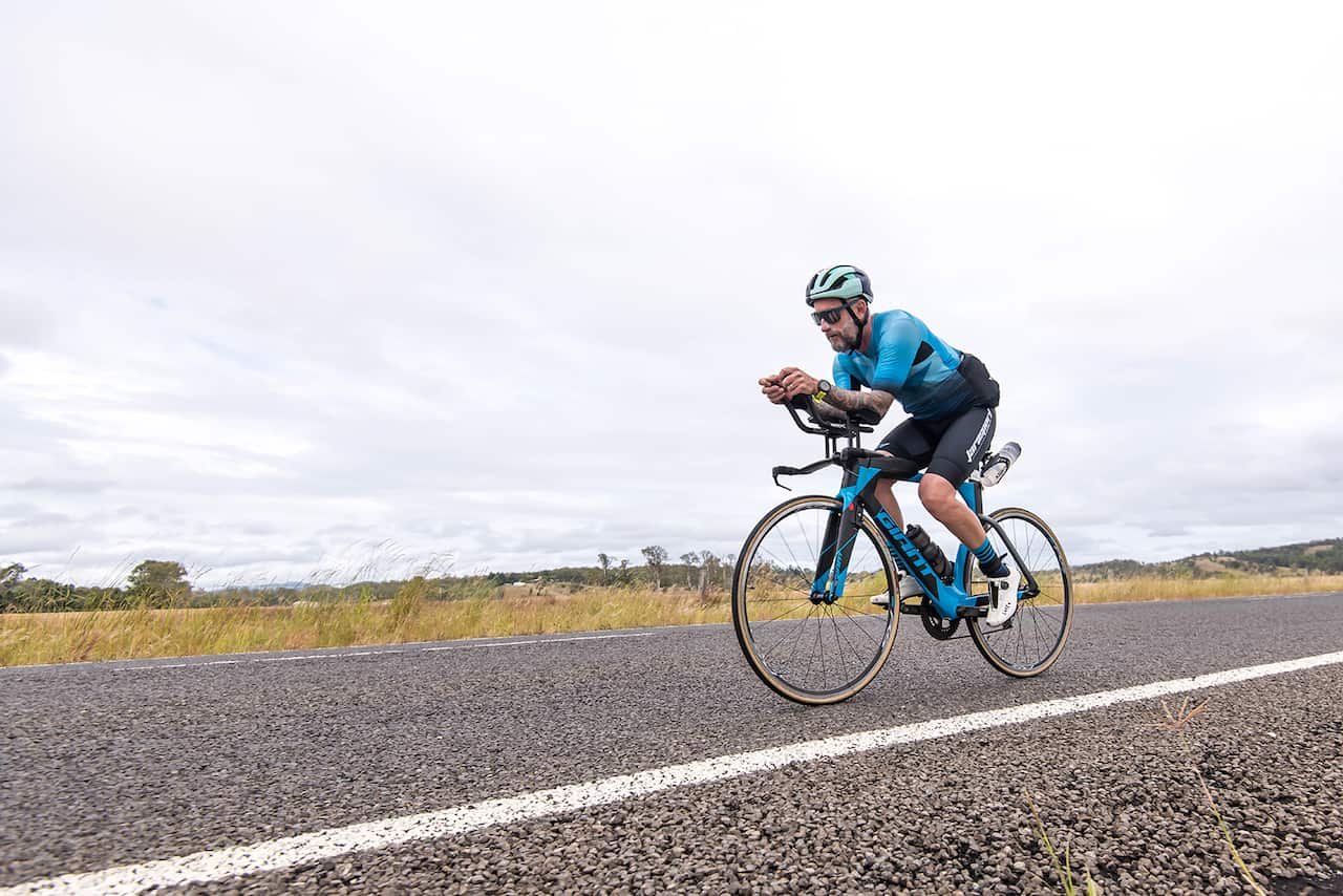 A man wearing an aqua-coloured cycling shirt and black cycling shorts riding a bike on a country road