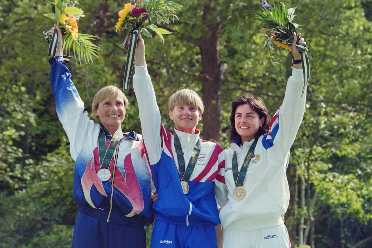 Three women wearing tracksuits and medals wave while holding bunches of flowers