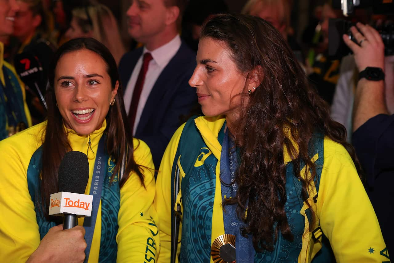 A woman watches as another woman speaks into a microphone. Both are wearing gold and green tracksuits.