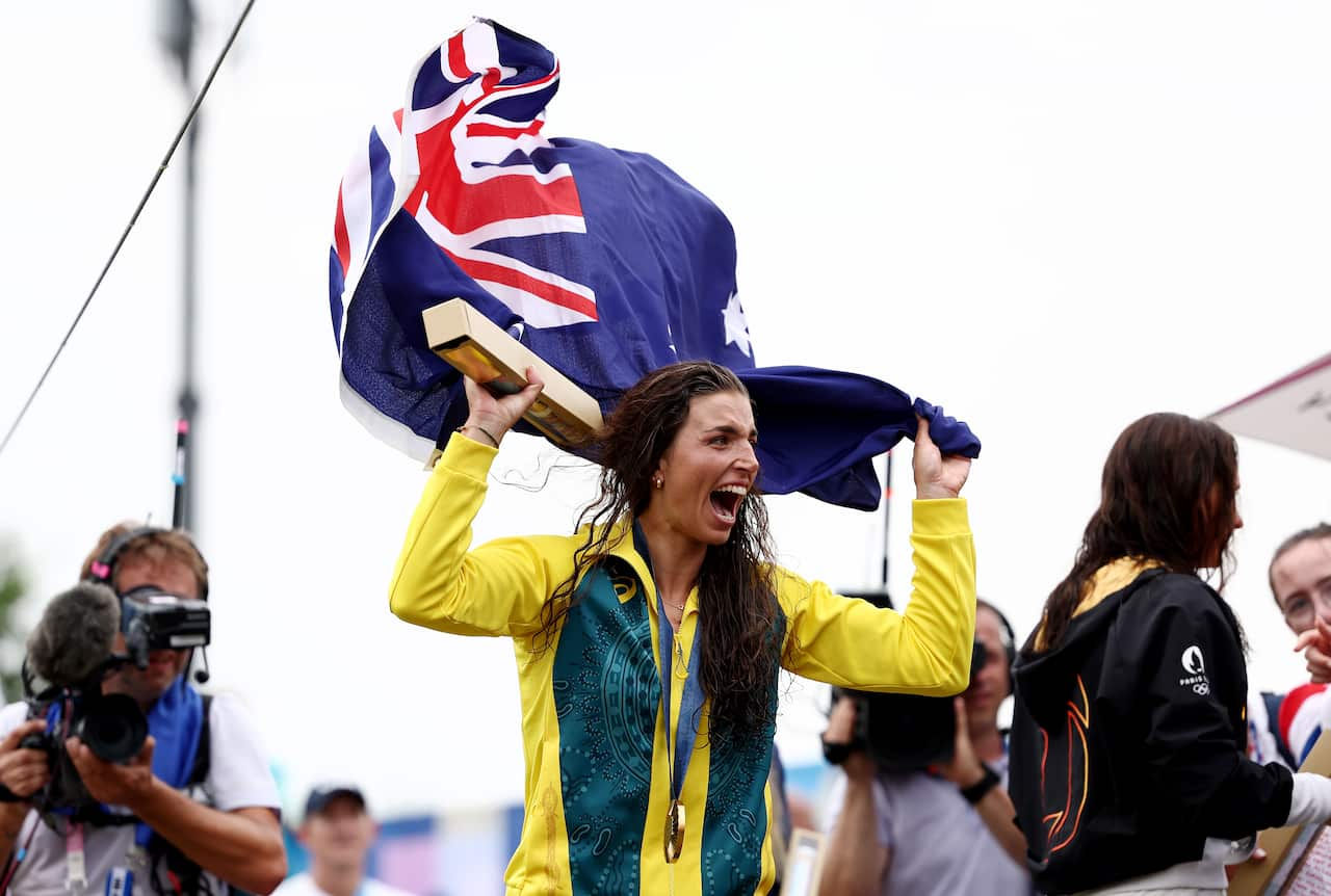 A woman wearing a green and yellow tracksuit smiles and waves an Australian flag over her head.