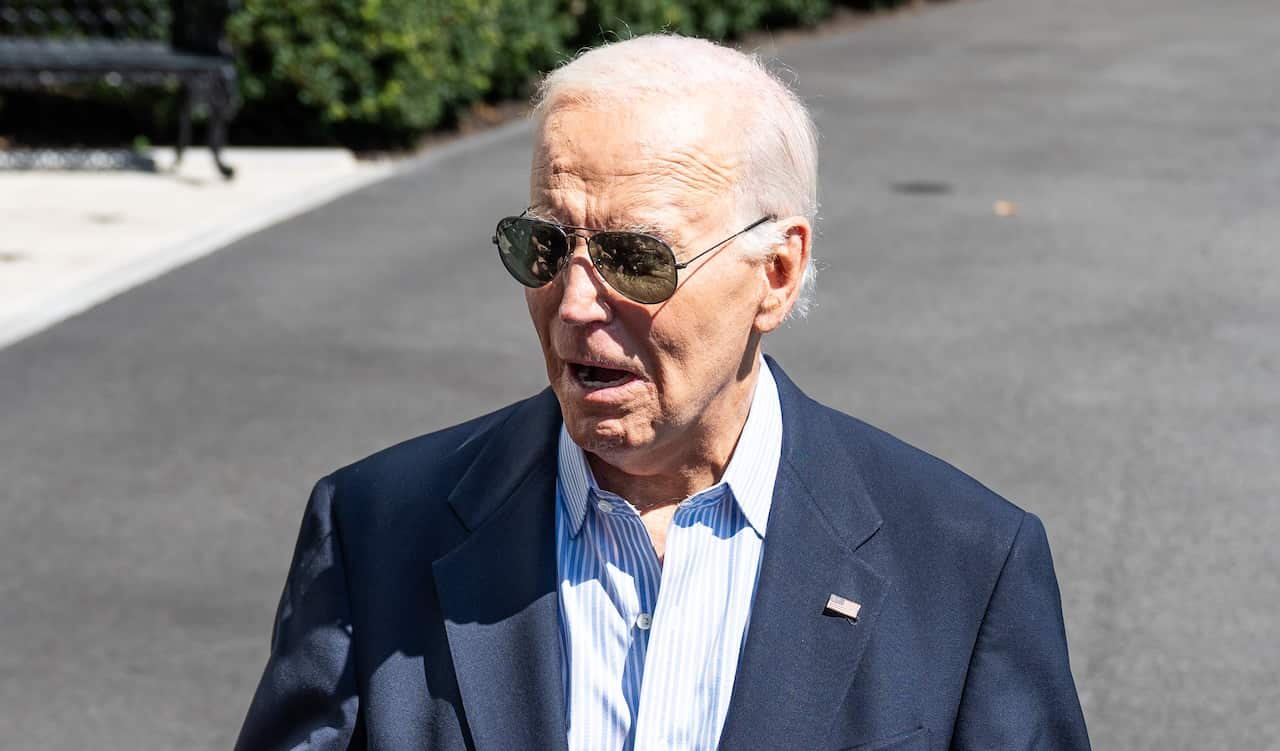 US President Joe Biden wearing a blue blazer with an American flag on it, blue and white shirt, and sunglasses.