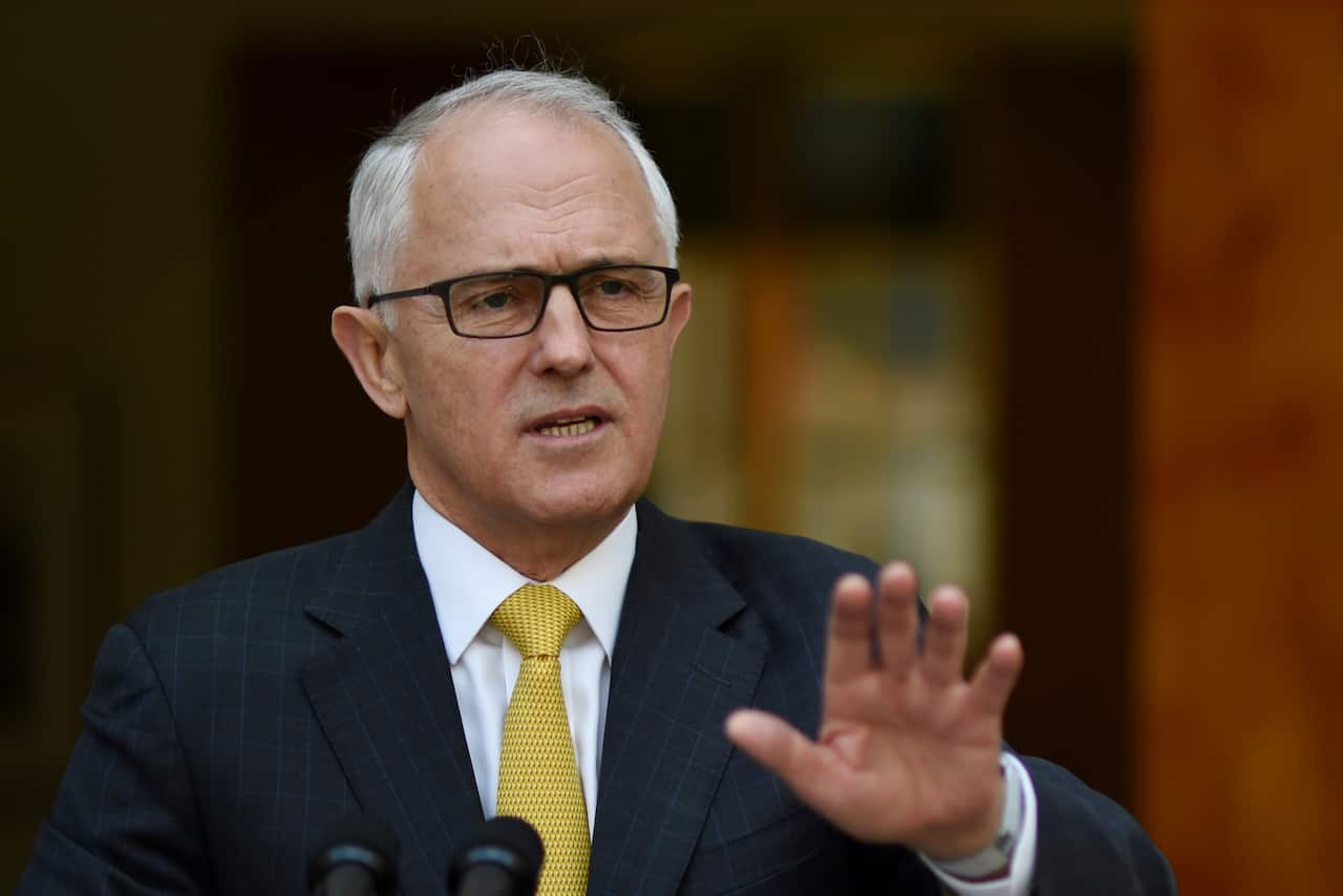 a man wearing glasses, a dark suit with a yellow tie, speaking in a formal setting. He is making a gesture with his hand.