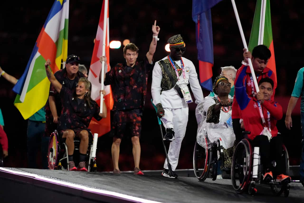 Athletes, including some in wheelchairs, and one using a crutch, come down a ramp. Some are holding flags