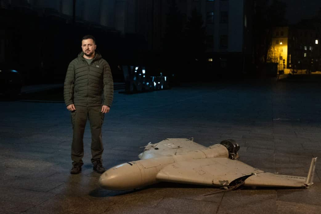 A man wearing a dark green puffer jacket stands next to a damaged drone aircraft