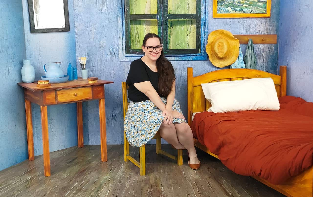 A young woman is smiling at the camera while sitting in a bedroom.