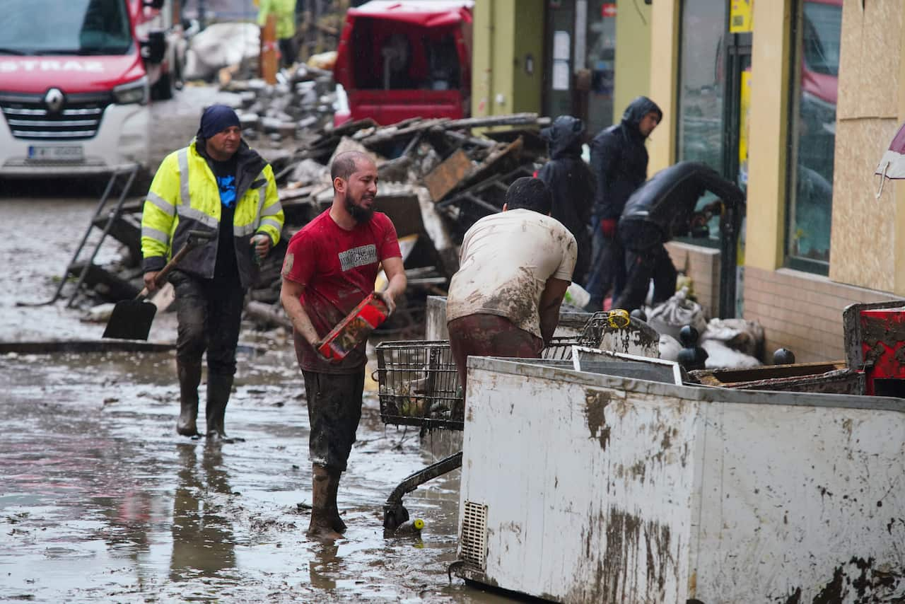 People walking in muddy street, two men carrying debris into shopping trolley. 