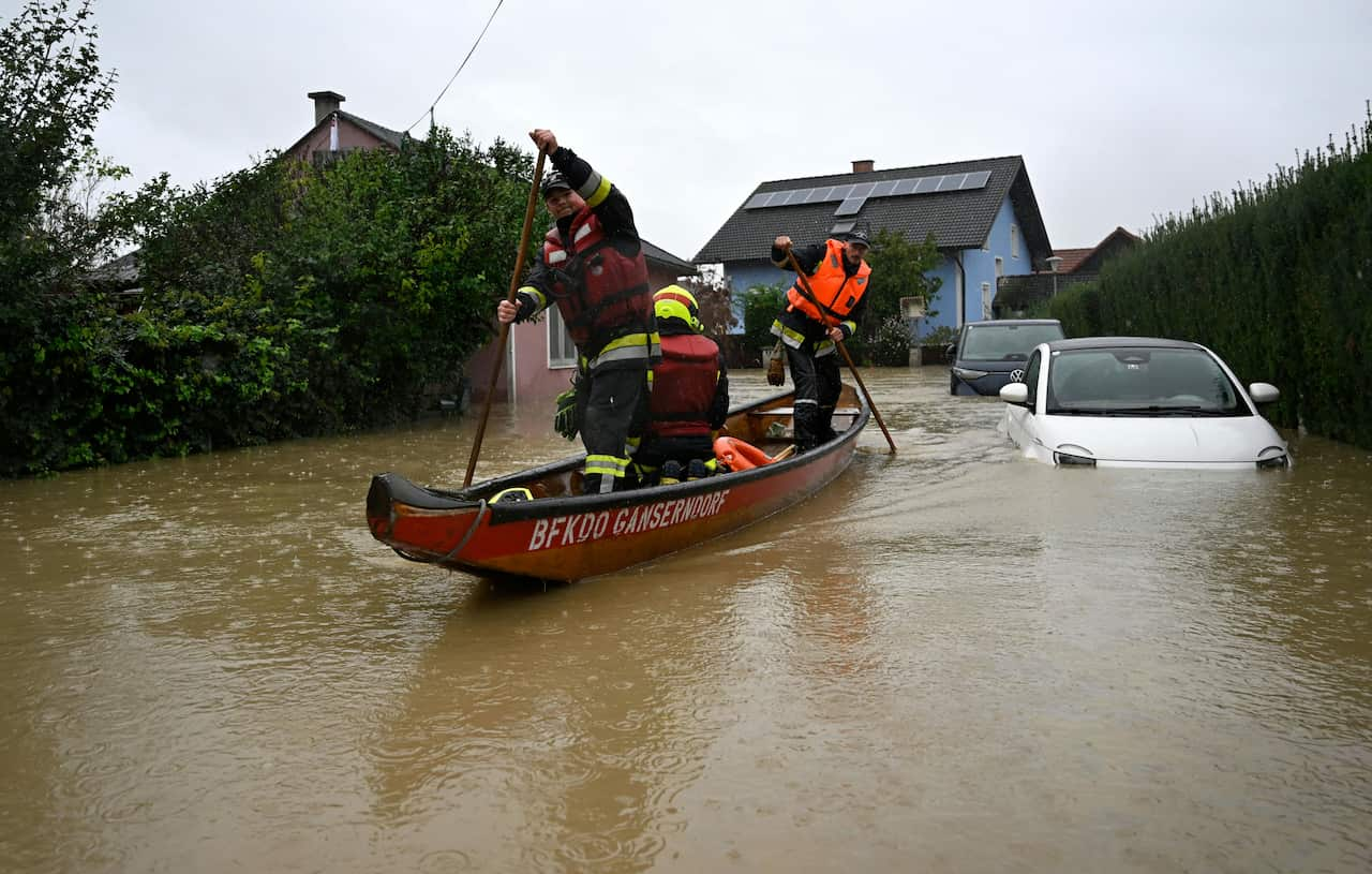 Men in high-vis paddling a small red boat through floodwaters, two cars submerged. 