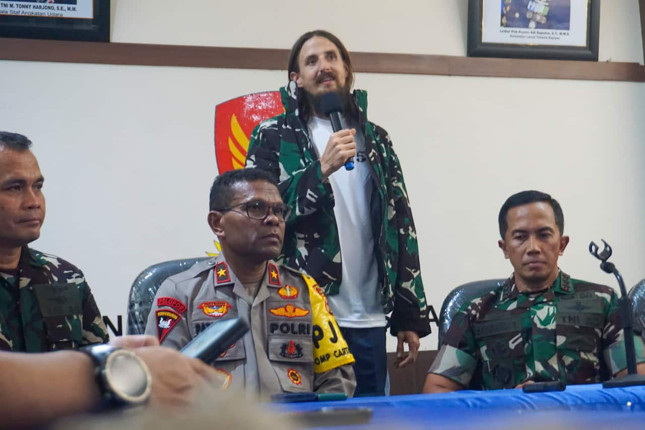 A man with a beard and long hair wearing an army jacket speaks into a microphone during a press conference. Several police and military personnel are seated in front of him. 