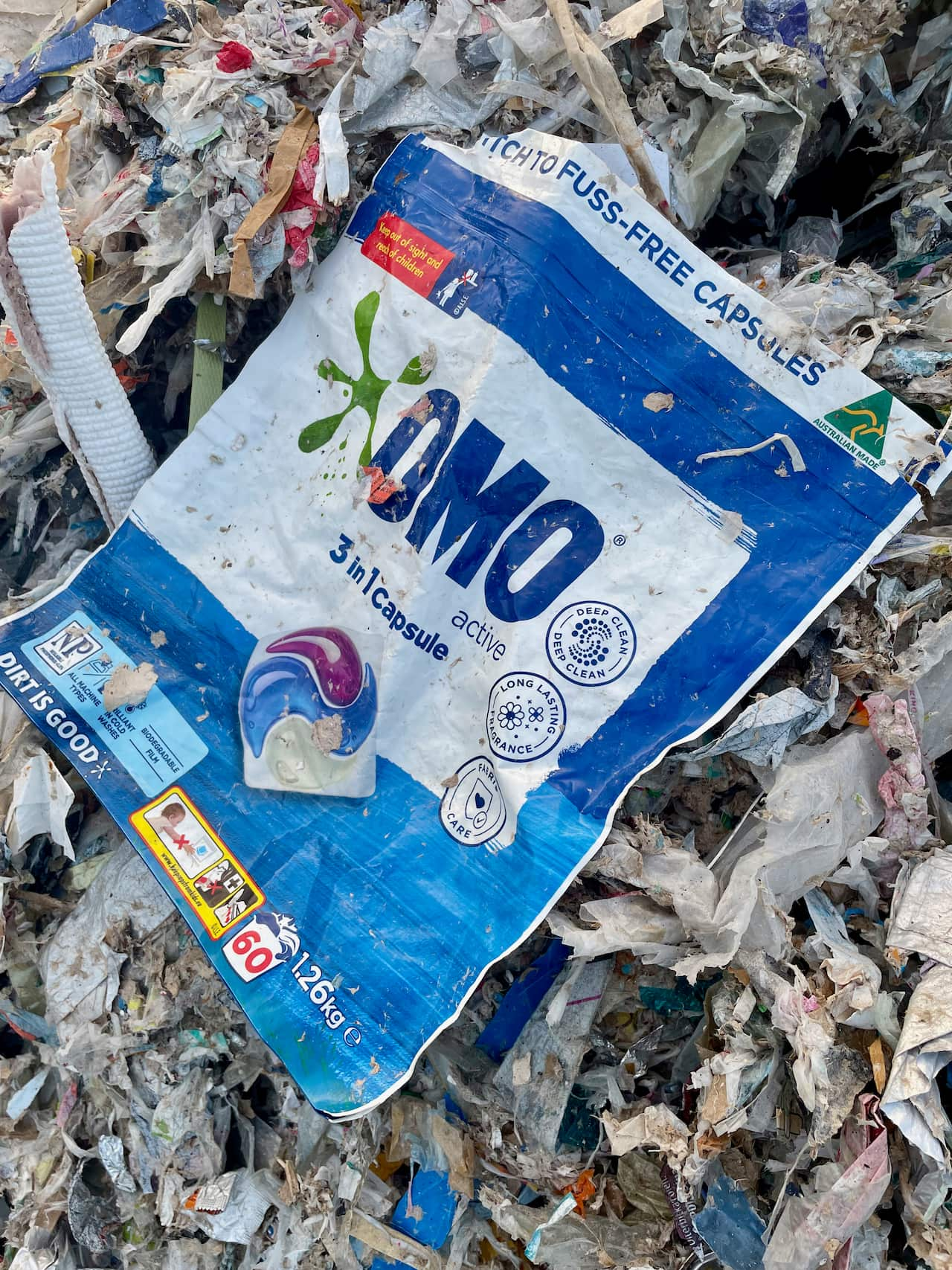 A discarded Australian-branded laundry capsule packet lies on a heap of waste paper and plastic.