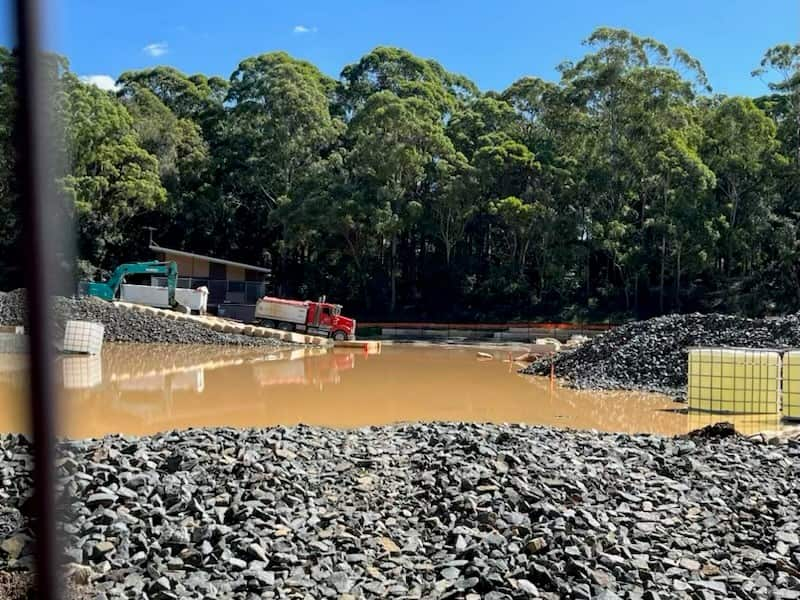 A construction site is flooded with murky water, with gravel in the foreground and truck machinery in the background.