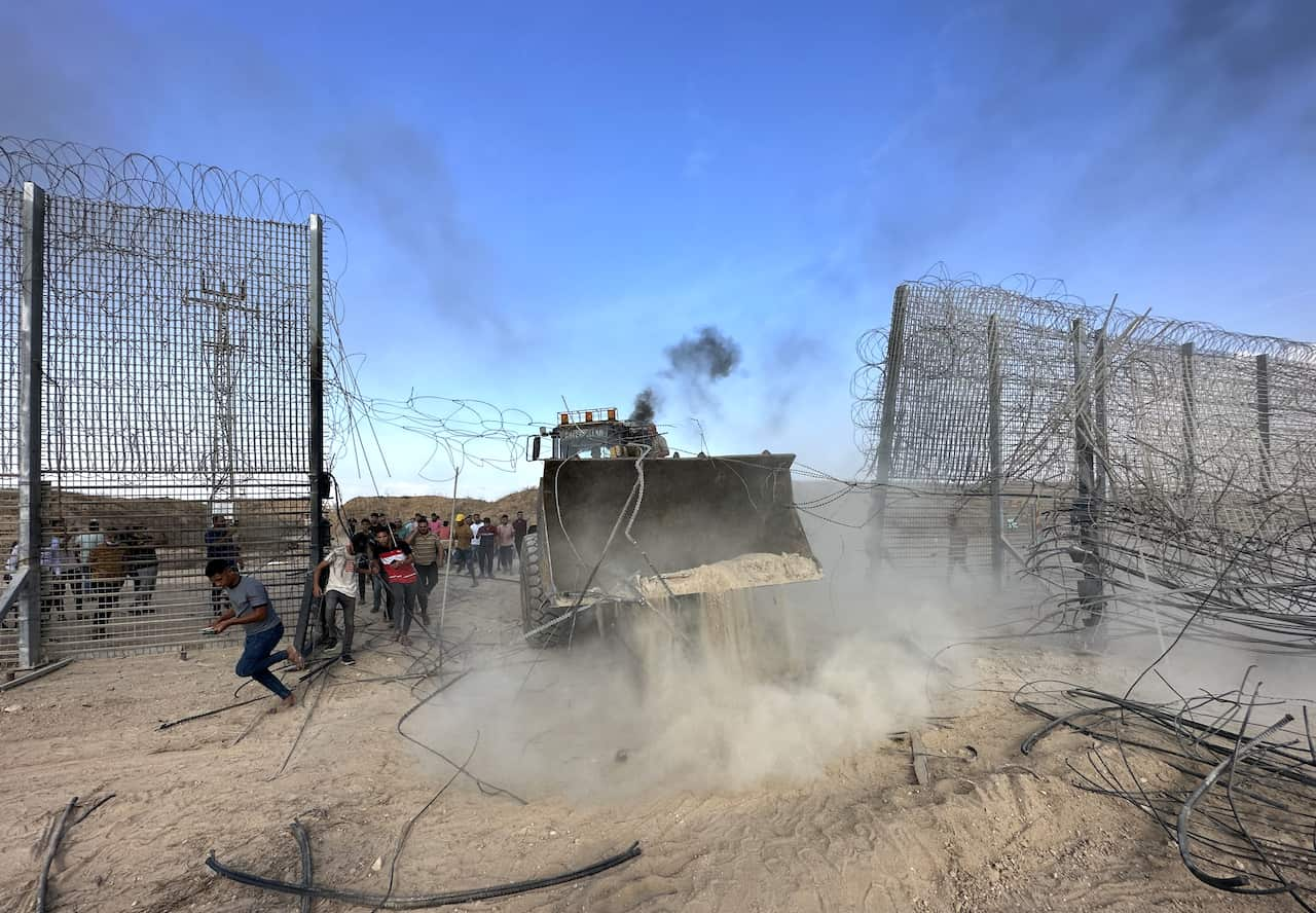 A bulldozer breaks through a wire fence as people dash through the gap.