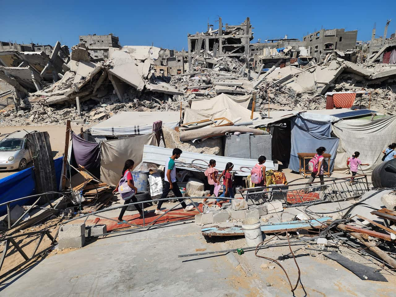 Children walk through rubble, past makeshift tents. There are destroyed buildings in the background.