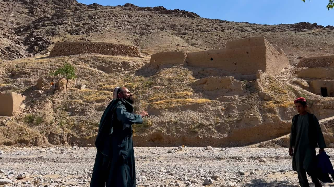 Two men in dark clothes and white and red hats stand before a sandy yellow cliff and structures. 