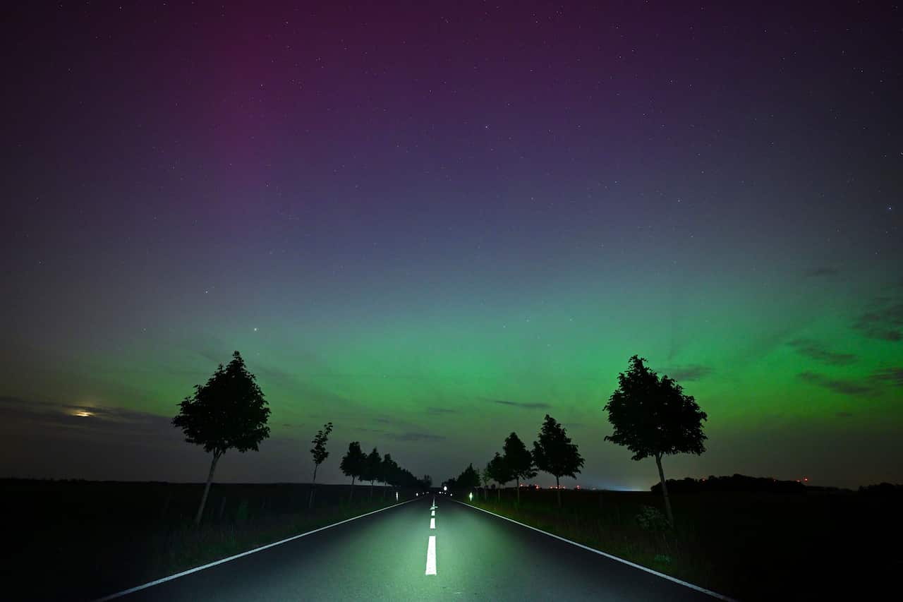Northern lights glow in the sky above a road with trees on either side.