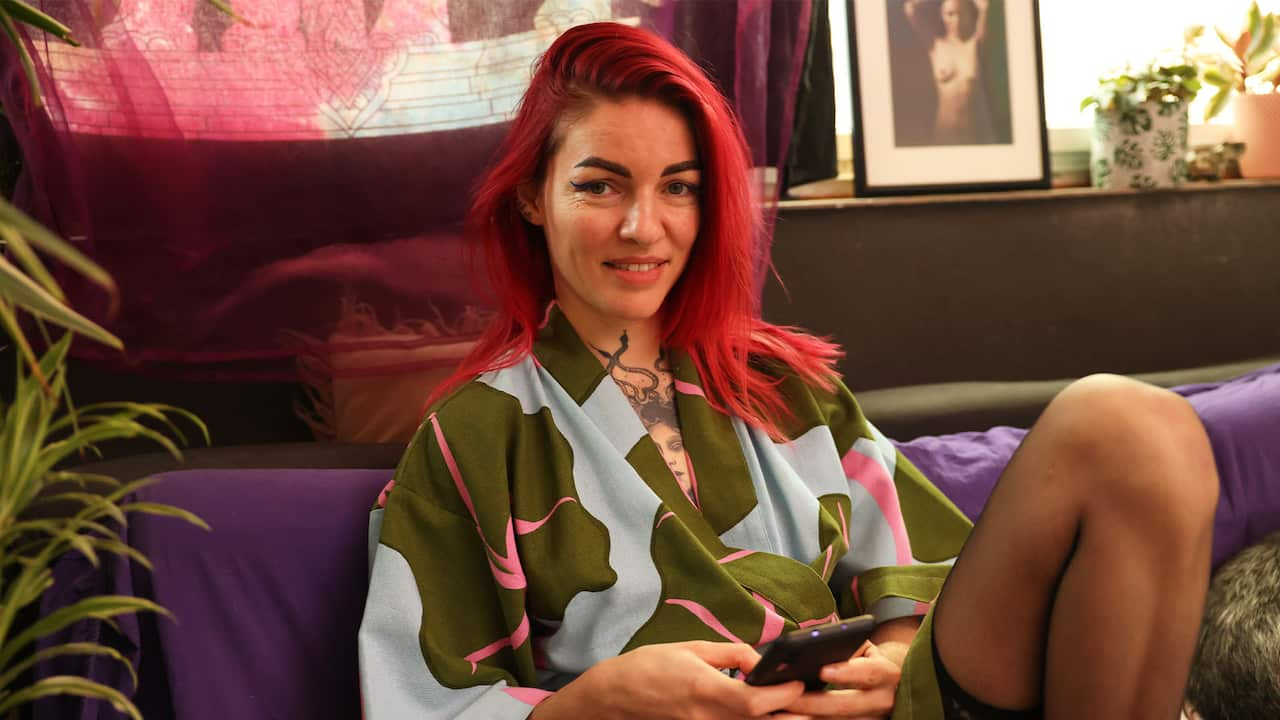 A woman with dyed red hair, wearing a blue and green robe and black pantyhose, leans back on a purple sofa. She is looking into the camera while holding her smartphone with both hands.