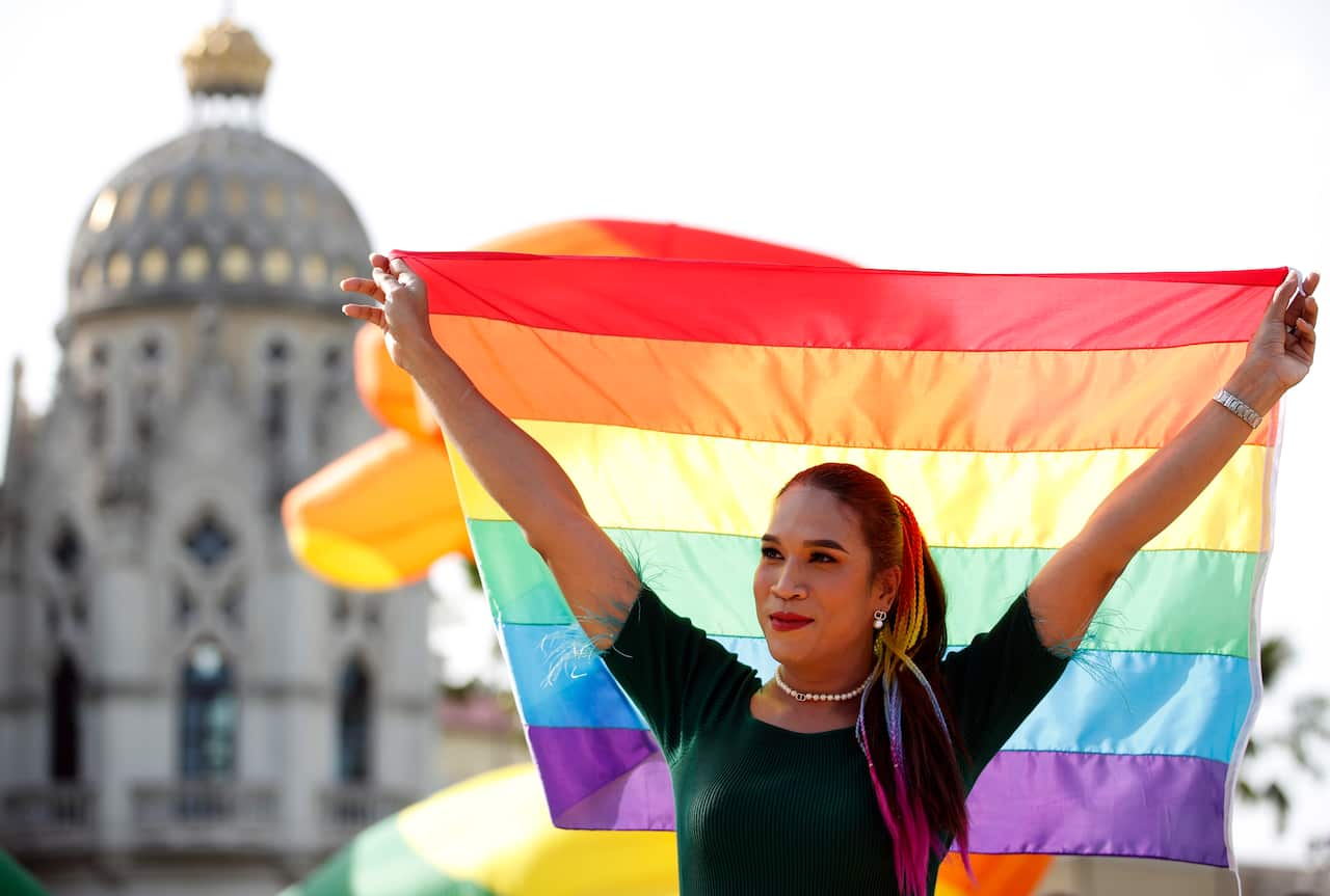 A person standing in front of a large building, smiling and holding up a rainbow flag.