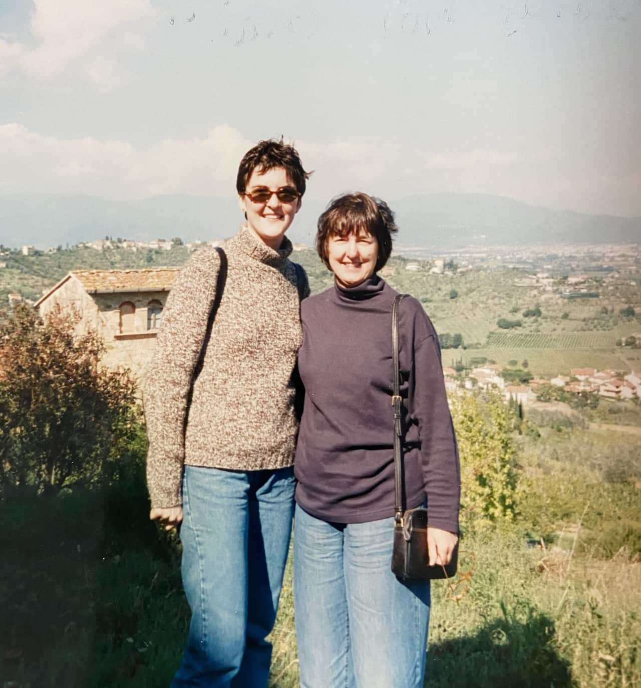 Two women stand together in the sunshine, smiling at the camera. It looks like they're on holiday.