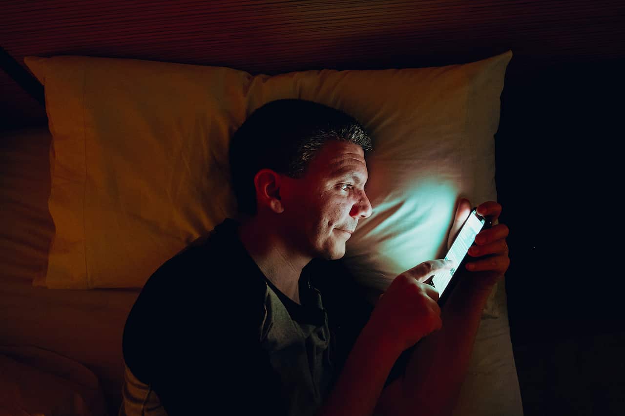 A man lying in bed looking at his mobile phone. A light is being emitted from the phone.