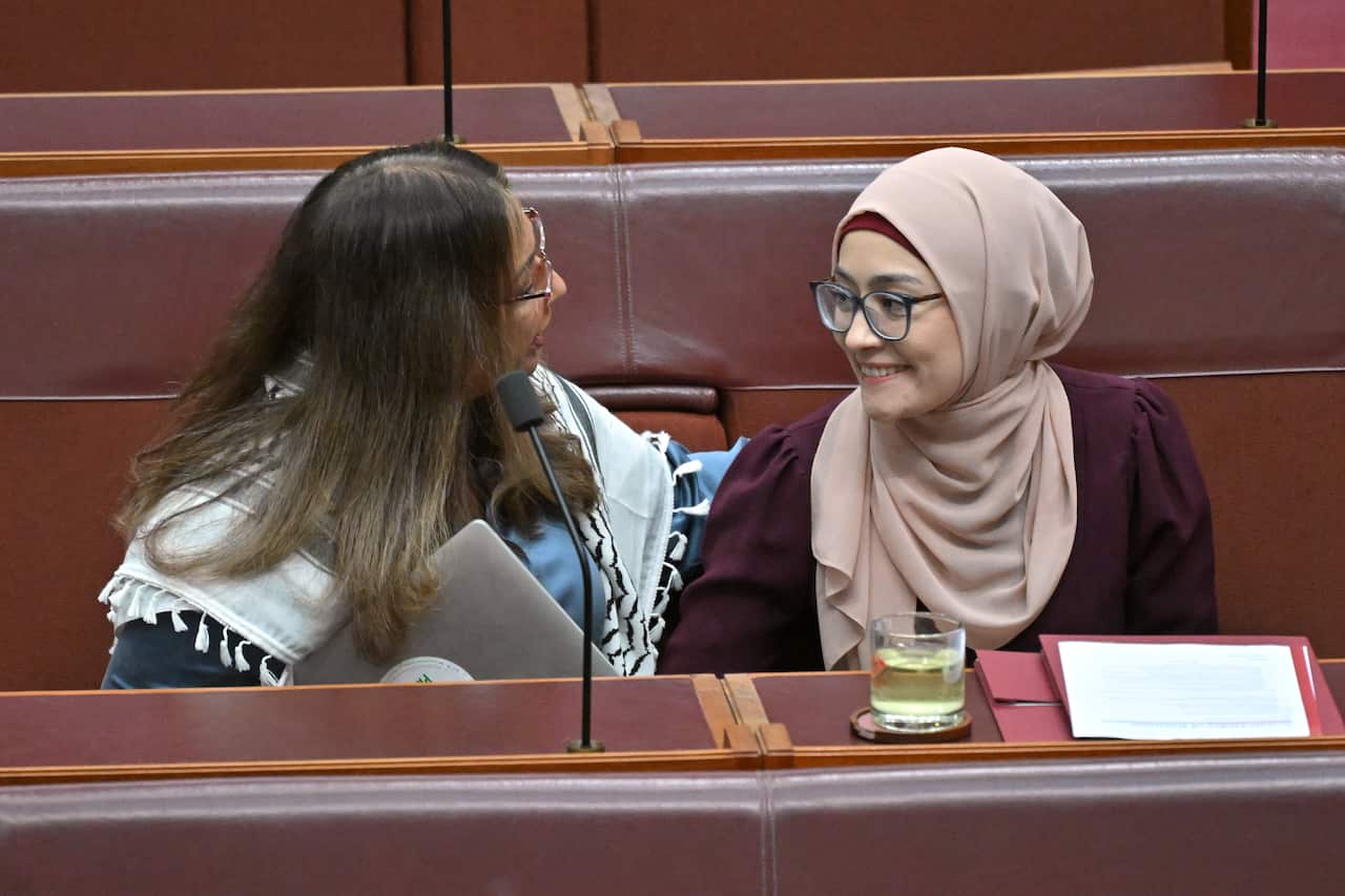 A woman wearing a keffiyeh places her arm on the shoulders of a younger woman wearing a headscarf. Both are sitting on red senate benches. 