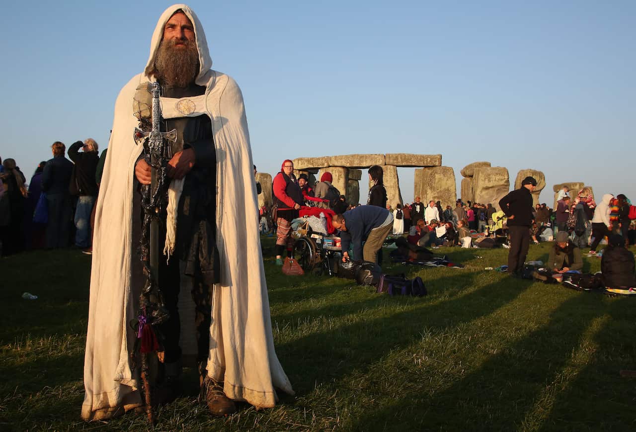 A man dressed as a druid stands in front of a group of people as the sun rises near Stonehenge.