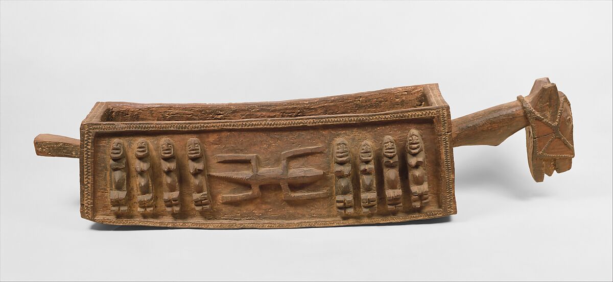 A two-metre long long wooden vessel with eight people carved into the side, four on each side separated by a carving of a lizard. The vessel has two handles on either side, one is in the shape of a horses head.