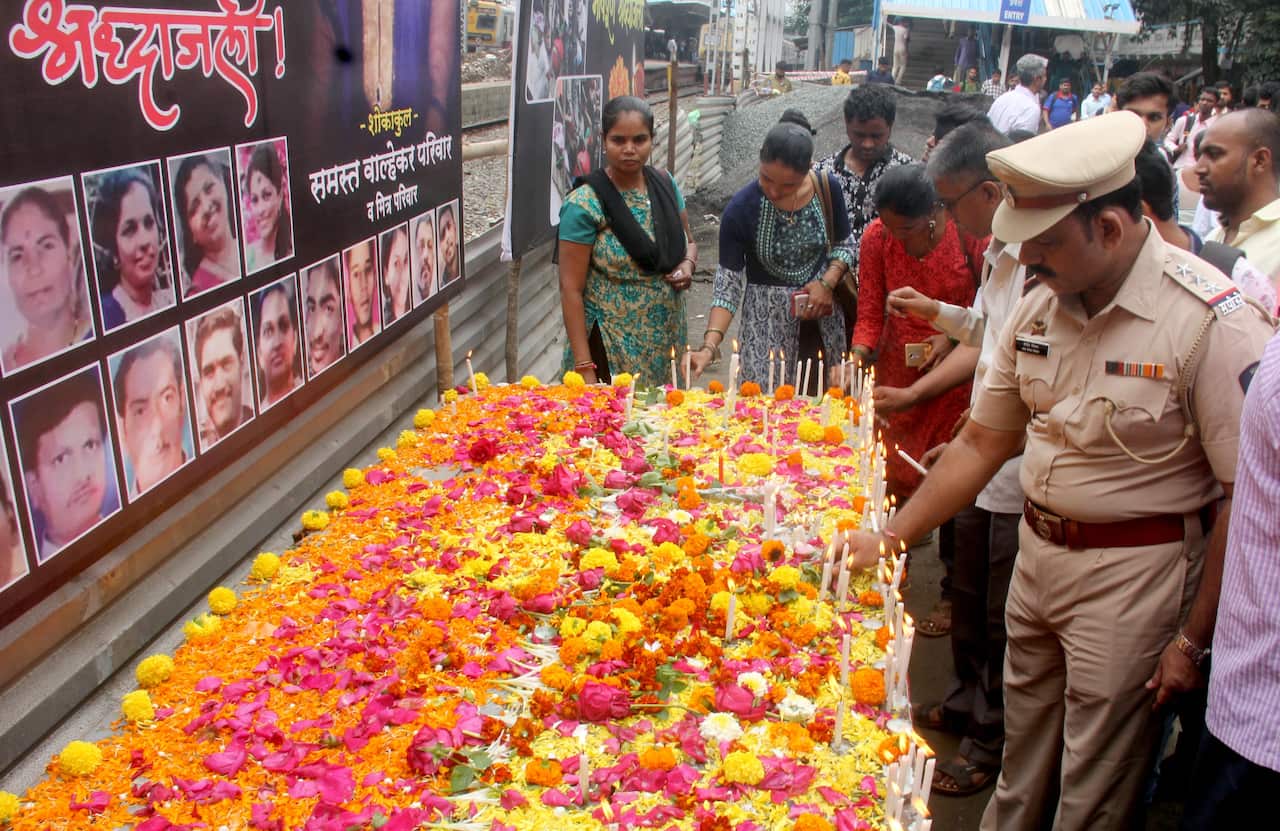 A group of people paying tribute to victims, whose faces are on a backdrop, with colourful flowers underneath them.