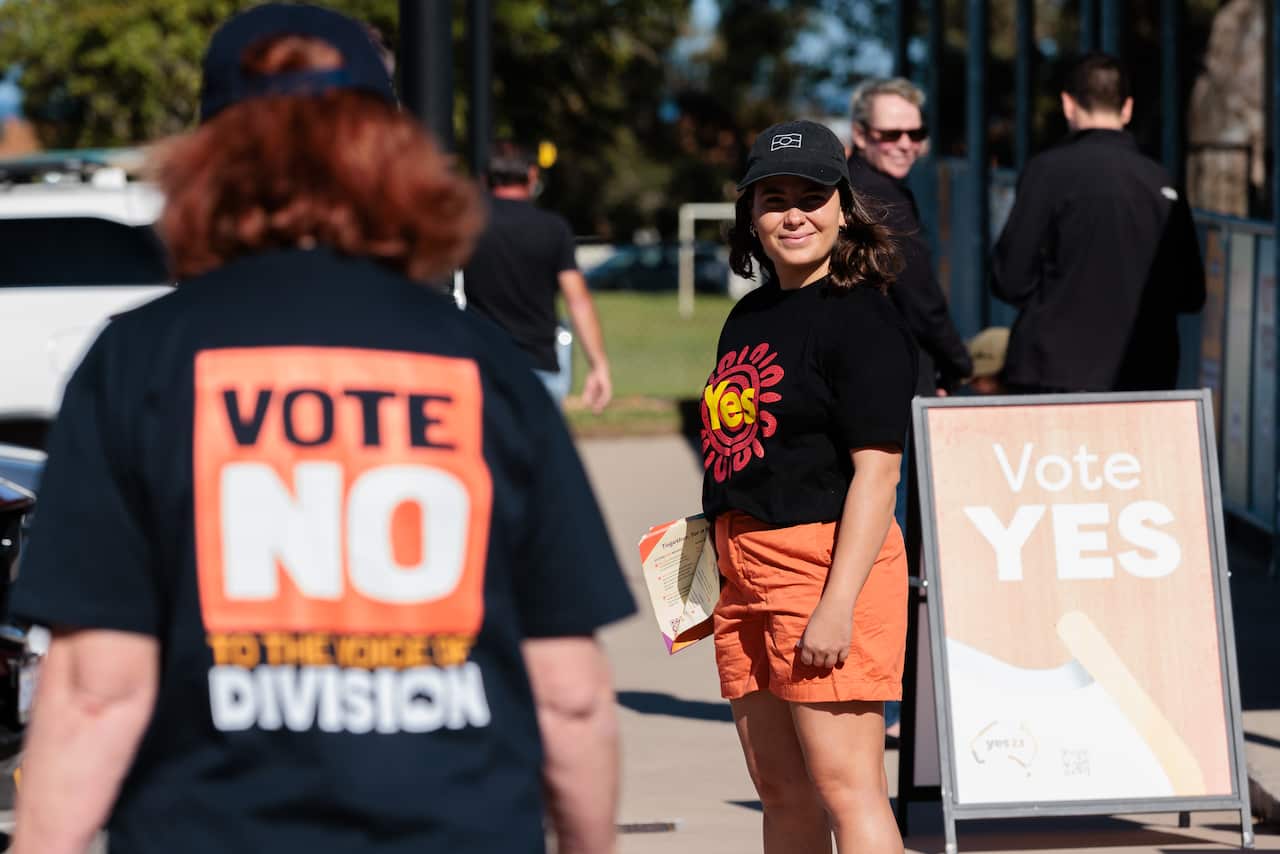 A person wearing a black cap stands beside a 'Vote Yes' sign with another person wearing a 'Vote No' t-shirt walking on.