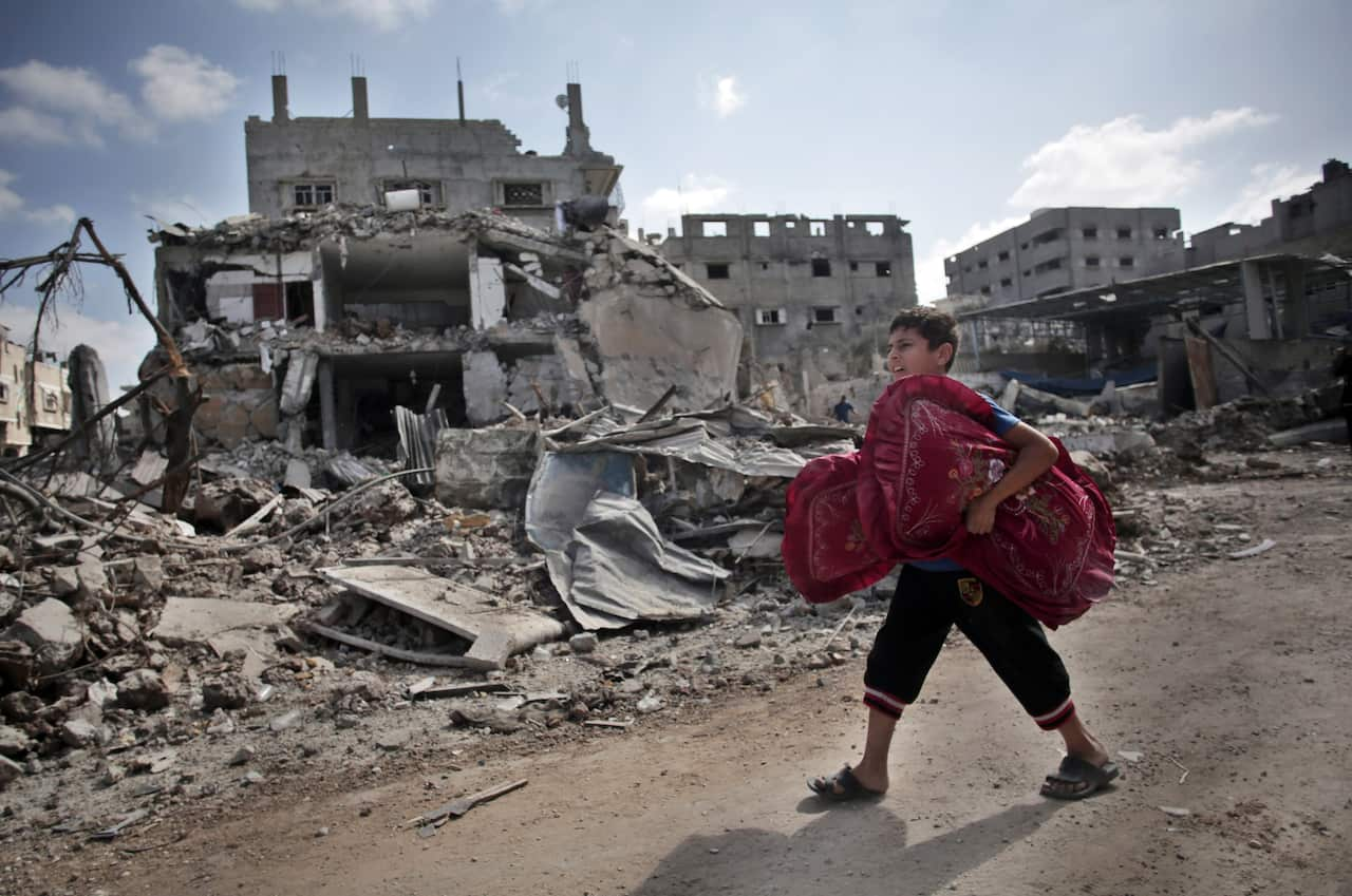 A Palestinian boy carries pillows as he passes by destroyed houses.