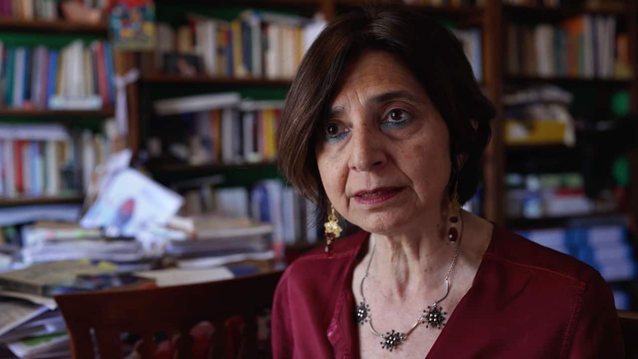 An older woman with short brown hair wearing a burgundy top and heavy jewellery standing in an office filled with books 