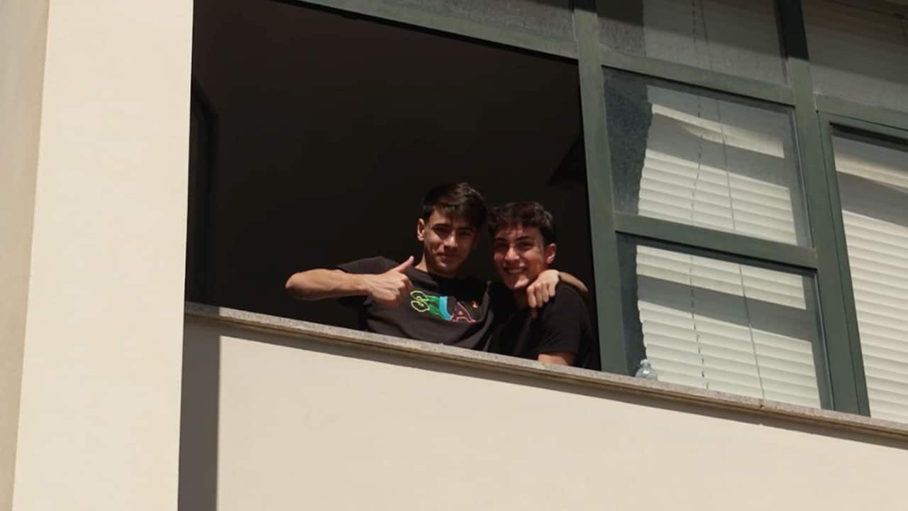 Two teenage boys seen in an open window. One of them is holding his left arm around the other's shoulder and is showing a thumb up.