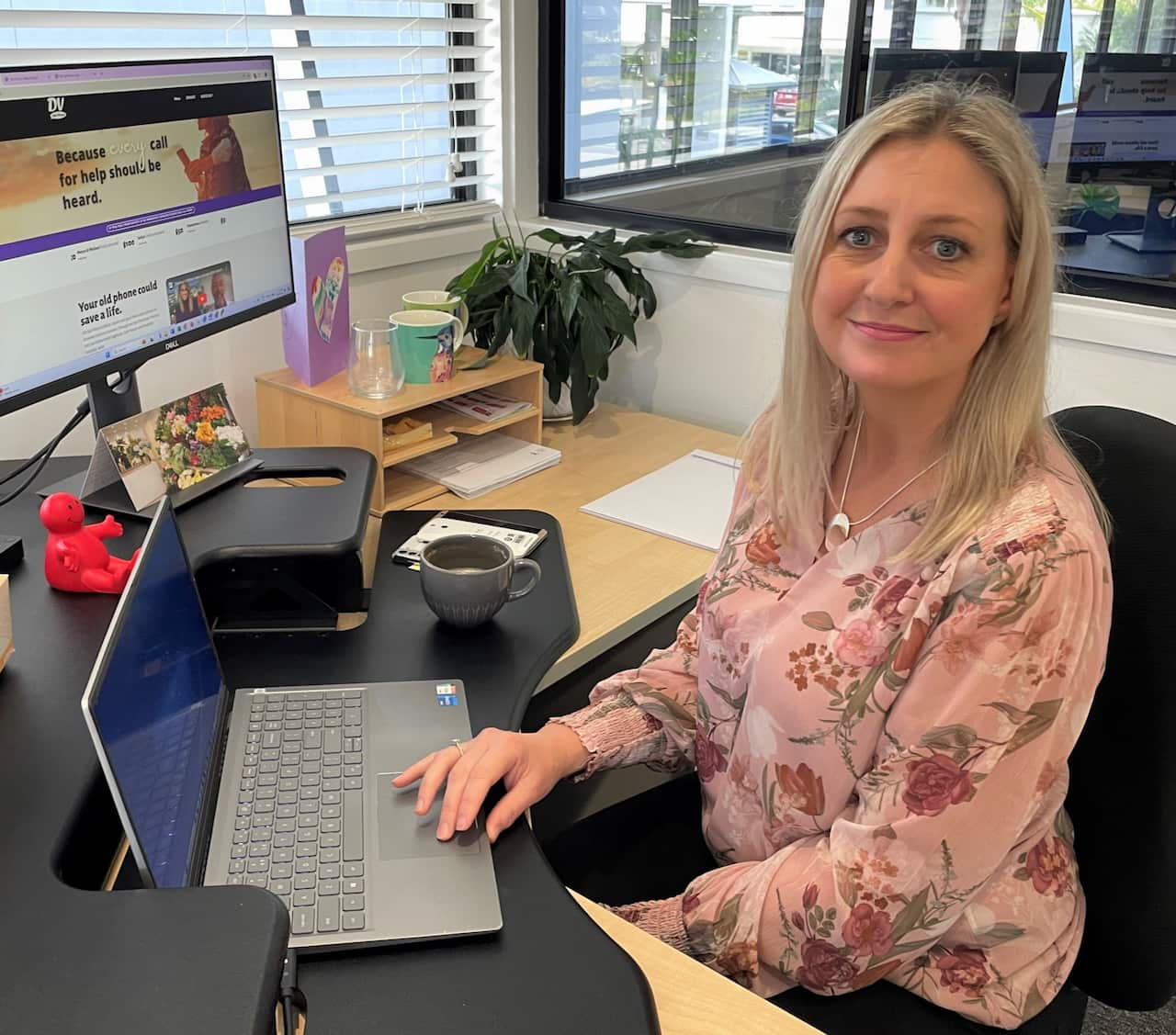 A woman in a floral dress sits at a computer in an office and smiles at camera. 