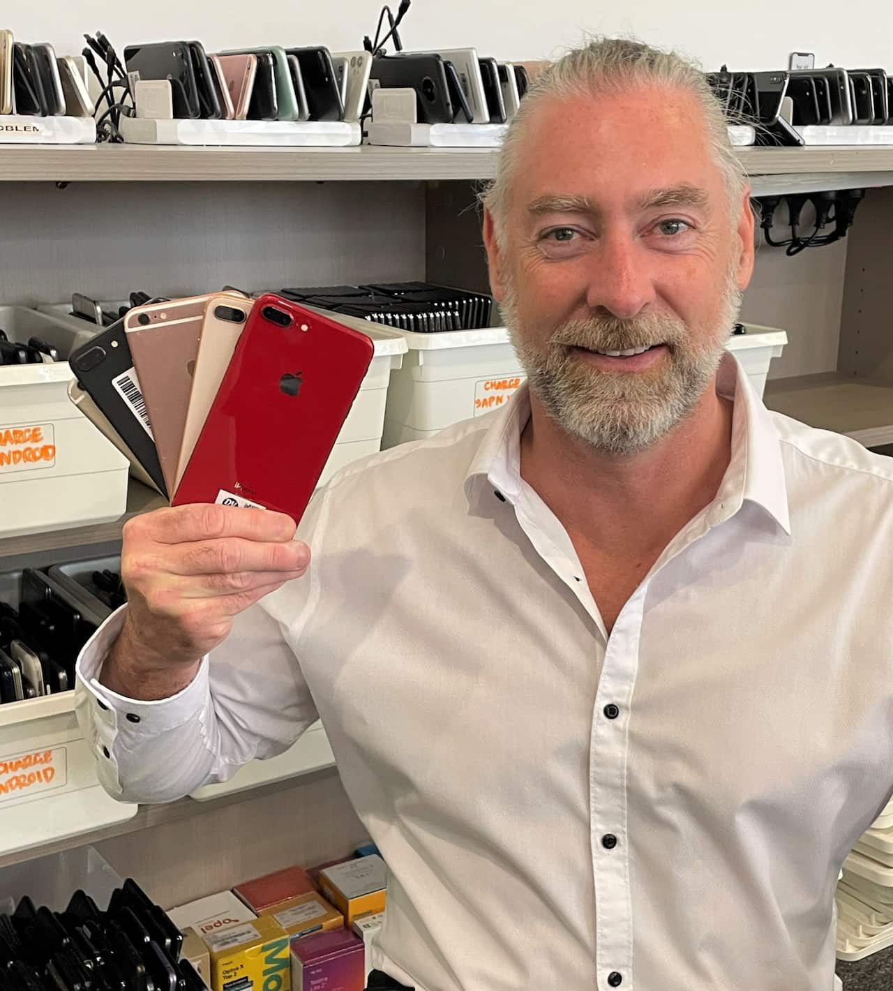 A man in a white shirt sits on a chair holding up mobile phones. 