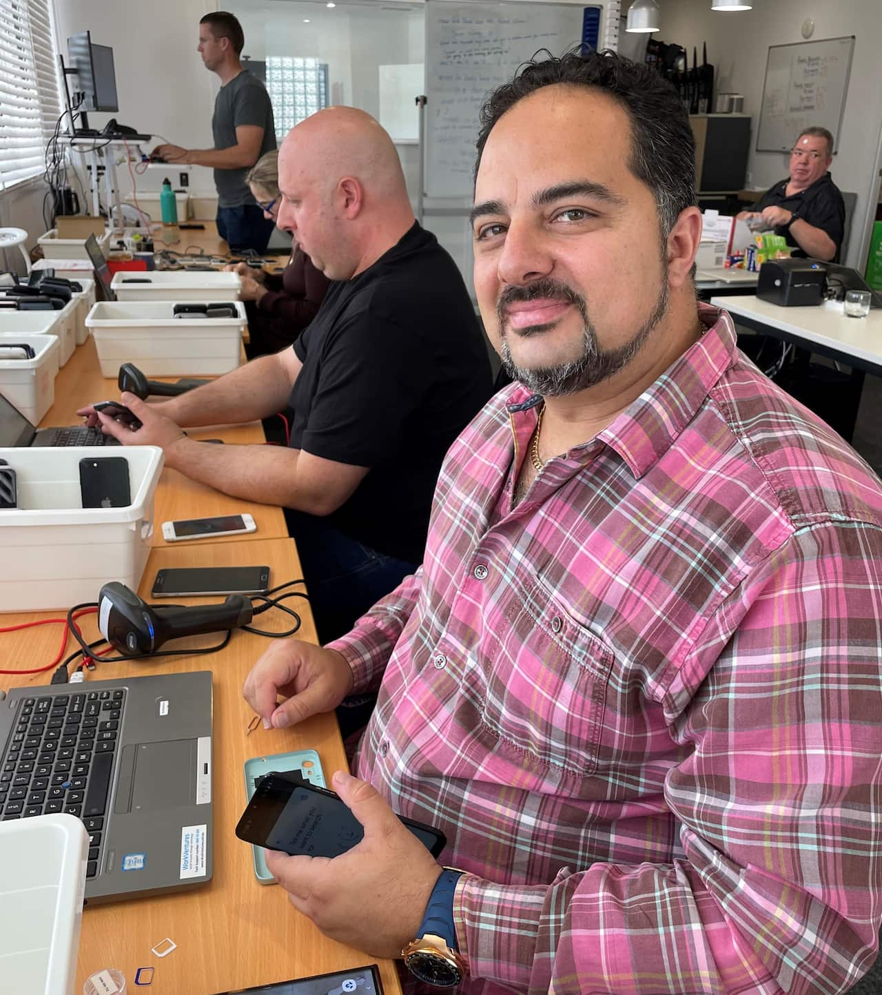 A man in a pink checked shirt sits at a bench working on a mobile phone.