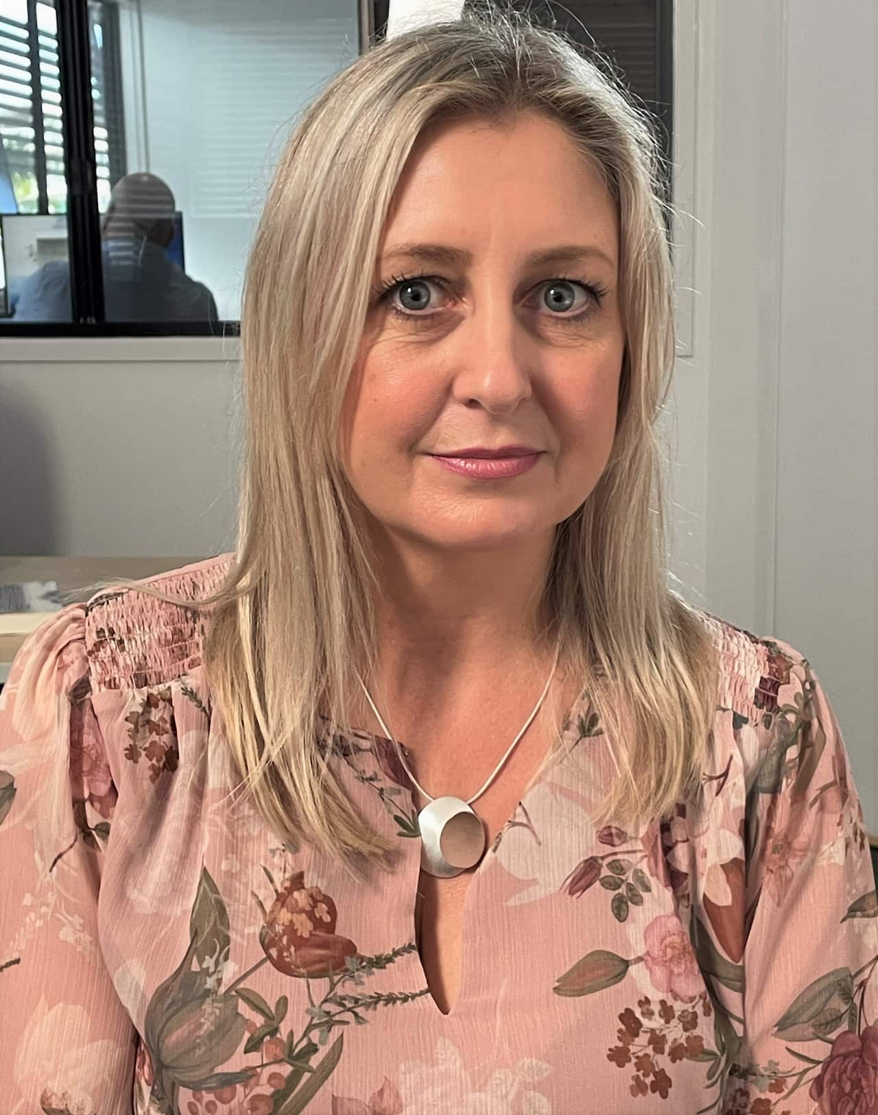 A woman in a pink floral dress faces the camera in an office.