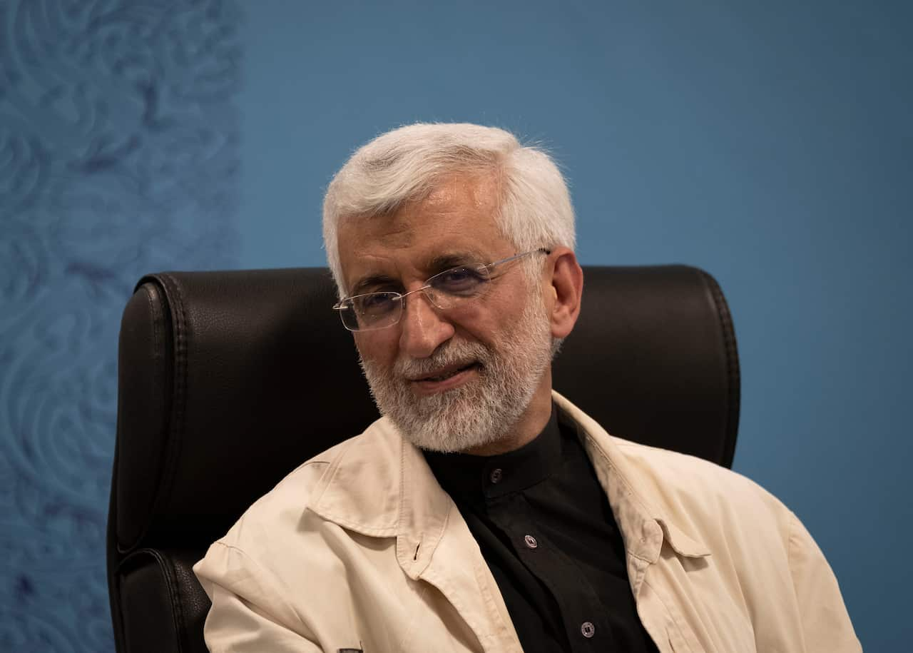 A man sitting on a black leather chair with a blue background behind him.