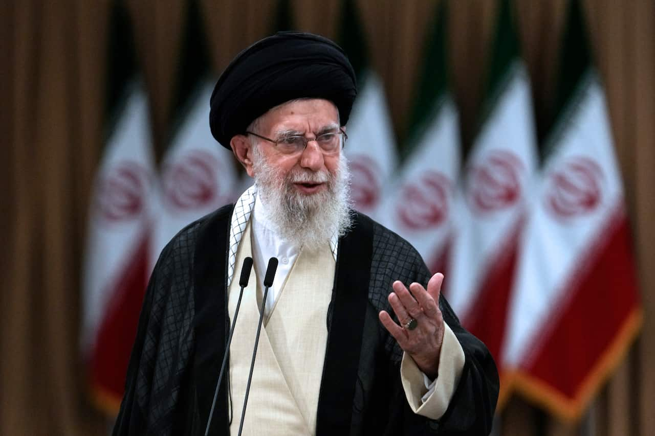 A man speaking into microphones in front of a row of Iranian flags.