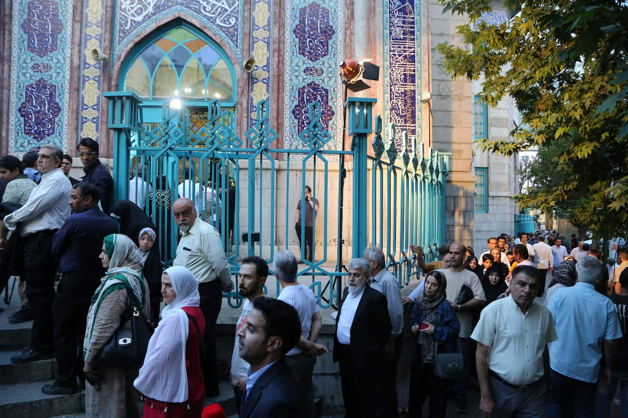 A large group of people standing outside a building in queue. There is a blue fence in front of the buildling.