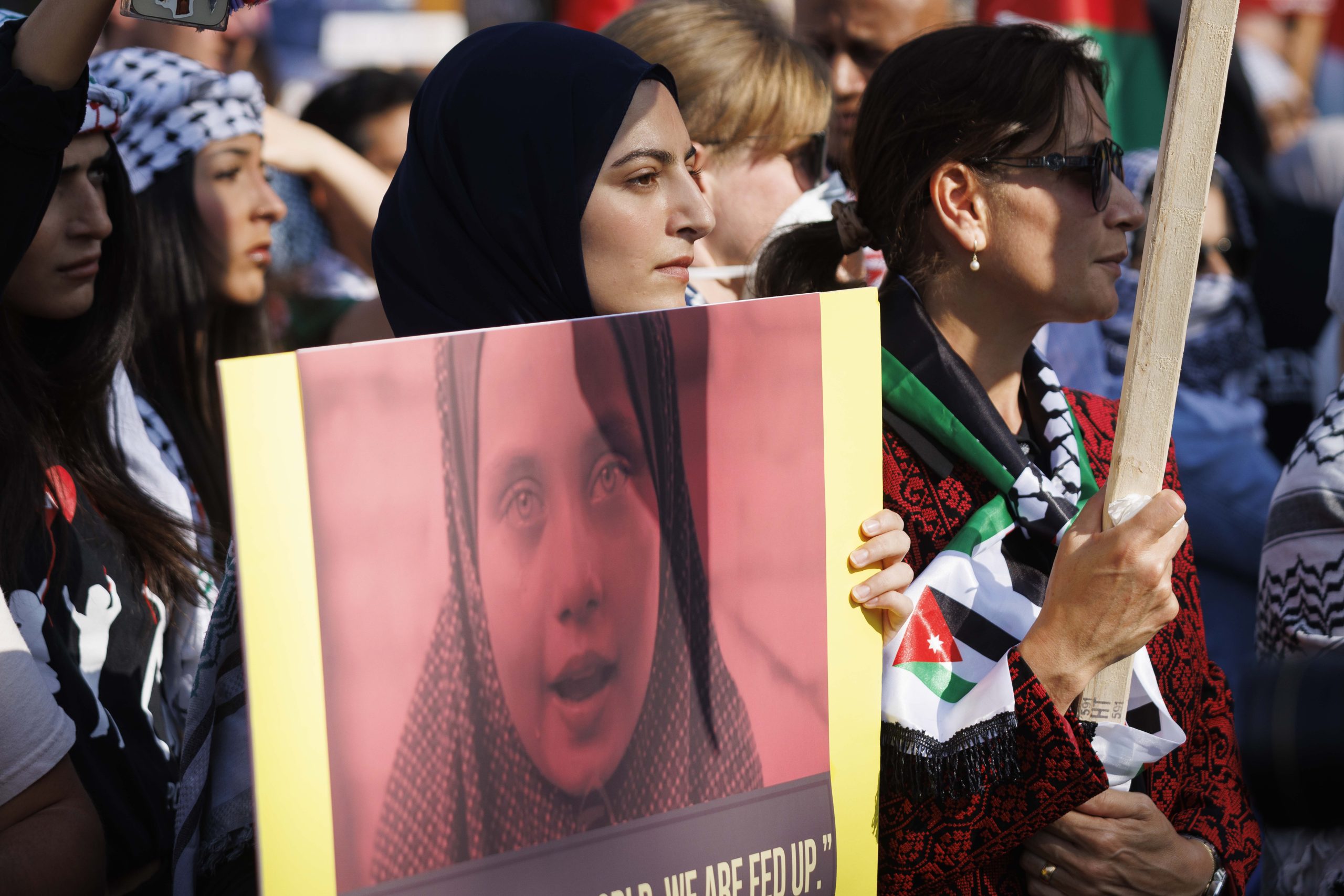 A female protestor wearing a hijab holds up an image of a child wearing a hijab and is surrounded by other protestors