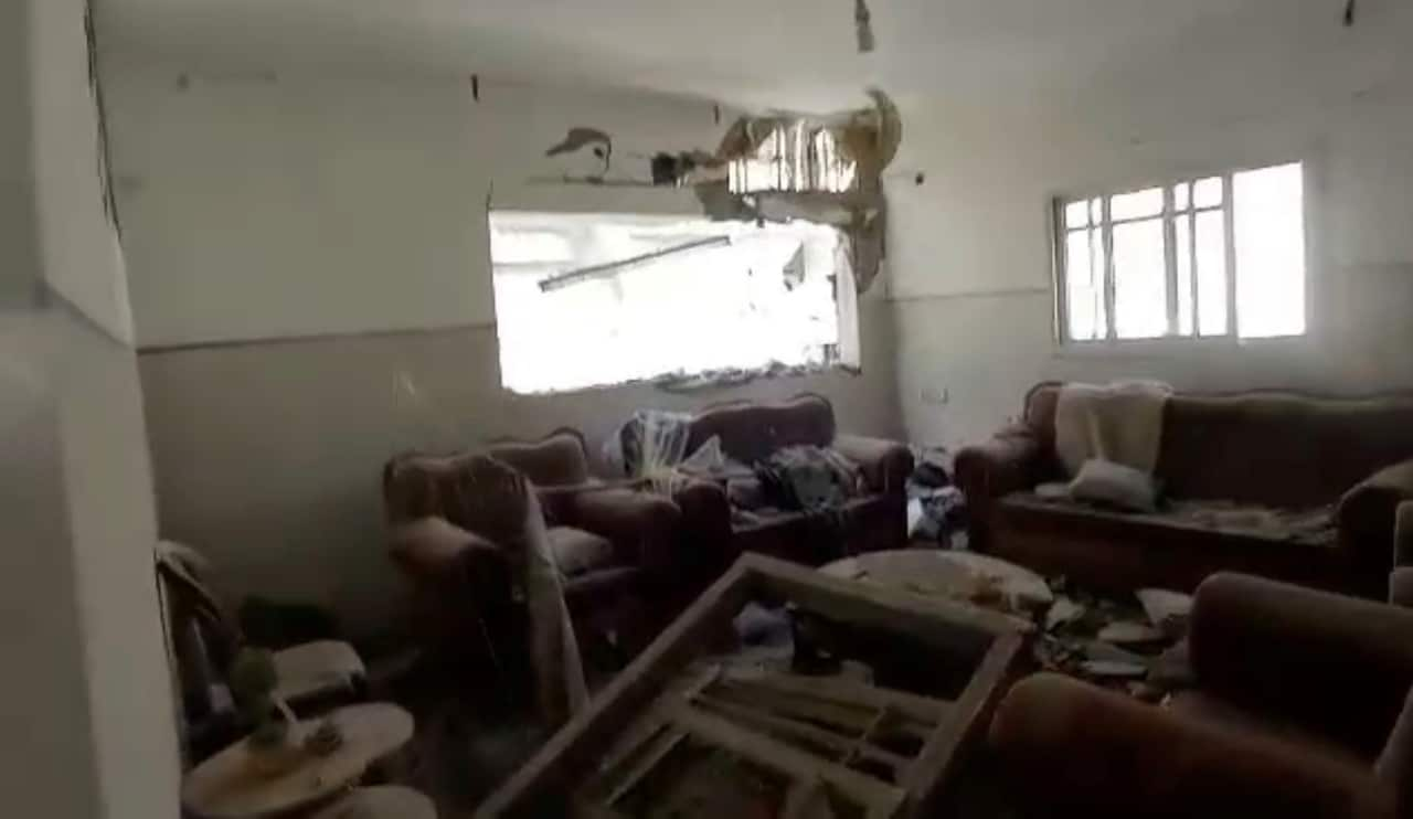 A loungeroom with red couches and armchairs scattered with debris, broken furniture and damage to the walls.