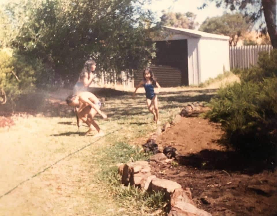 A young girl plays under a garden hose or sprinkler with her siblings.