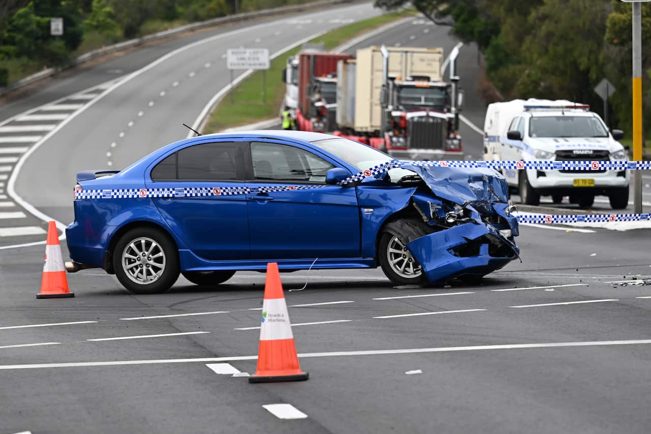 A blue car with a heavily dented front bumper sits in the middle of a road surrounded by police tape and orange safety cones. A police car is parked in the background behind it.