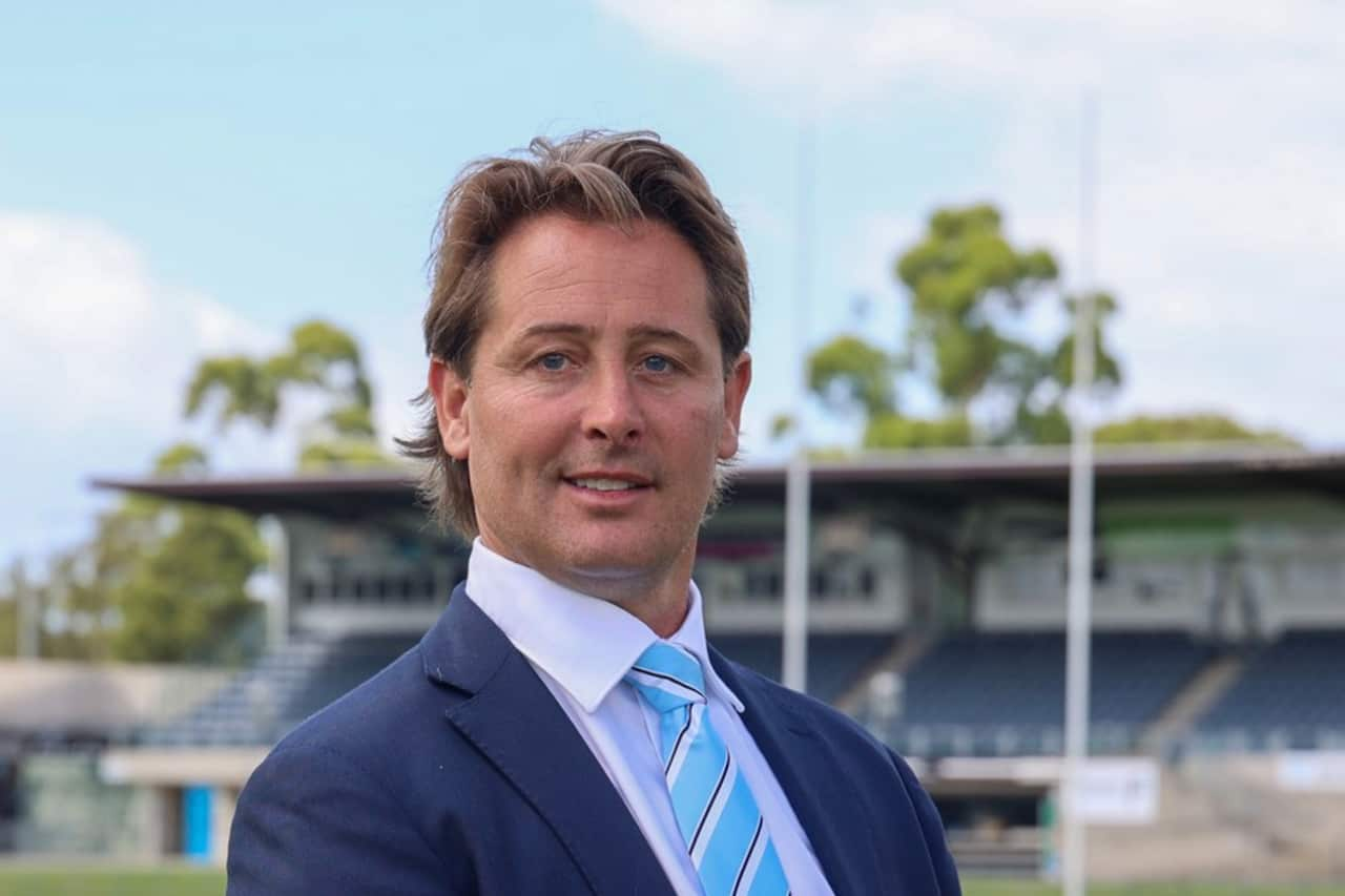 A man in a blue suit and tie looks forward with a slight smile on his face. He is standing in front of the stands of a sporting field. 