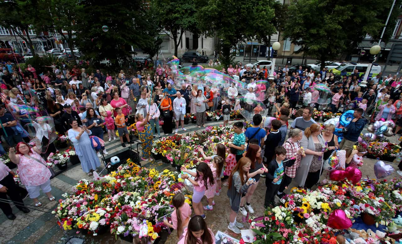 People attending an outdoor vigil. There are large bunches of flowers in the foreground