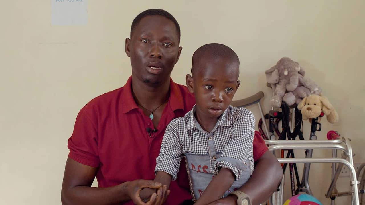 An African man wearing a red polo shirt holding a young boy on his lap. In the background, there are plush toys and walking crutches 